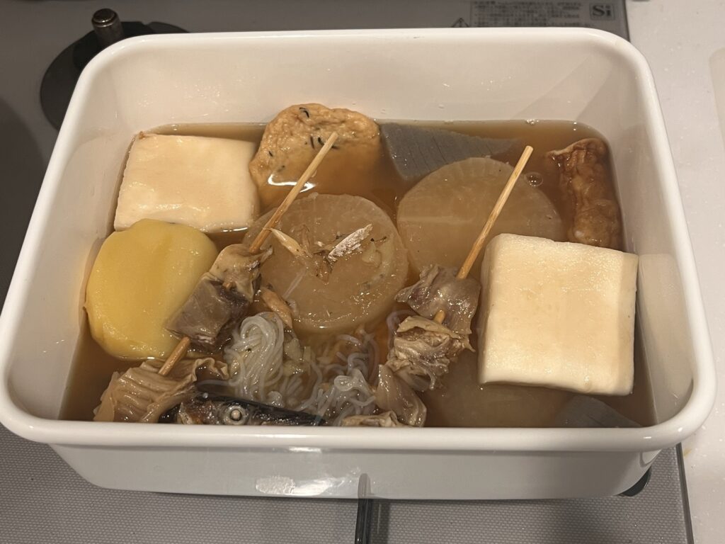白いホーロー容器に入ったおでん。大根やはんぺん、牛すじの串がだしに浸かっている